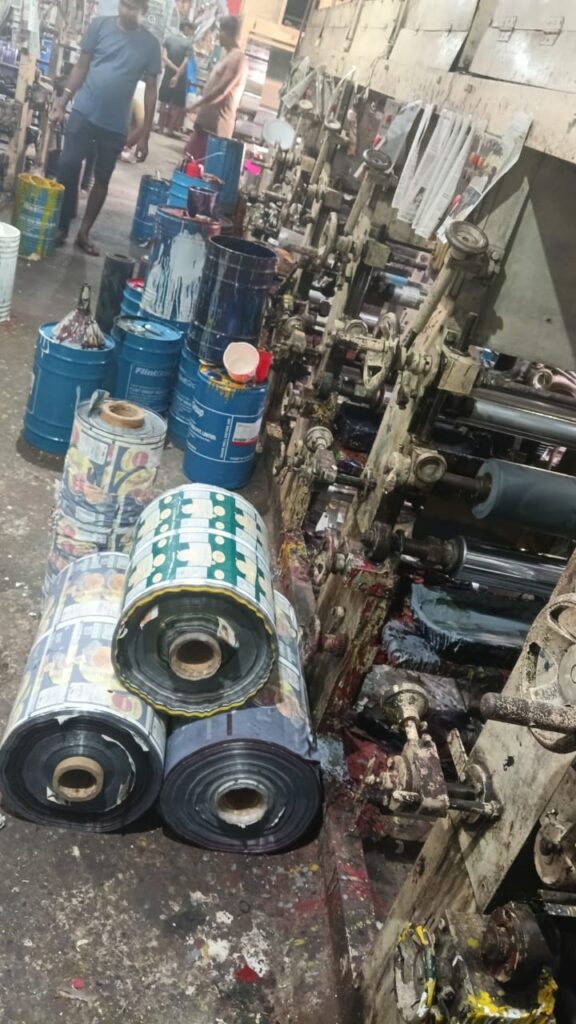 A busy printing facility with machinery, paint barrels, and rolls of printed material stacked on the floor, demonstrating the manufacturing process of packaging solutions.