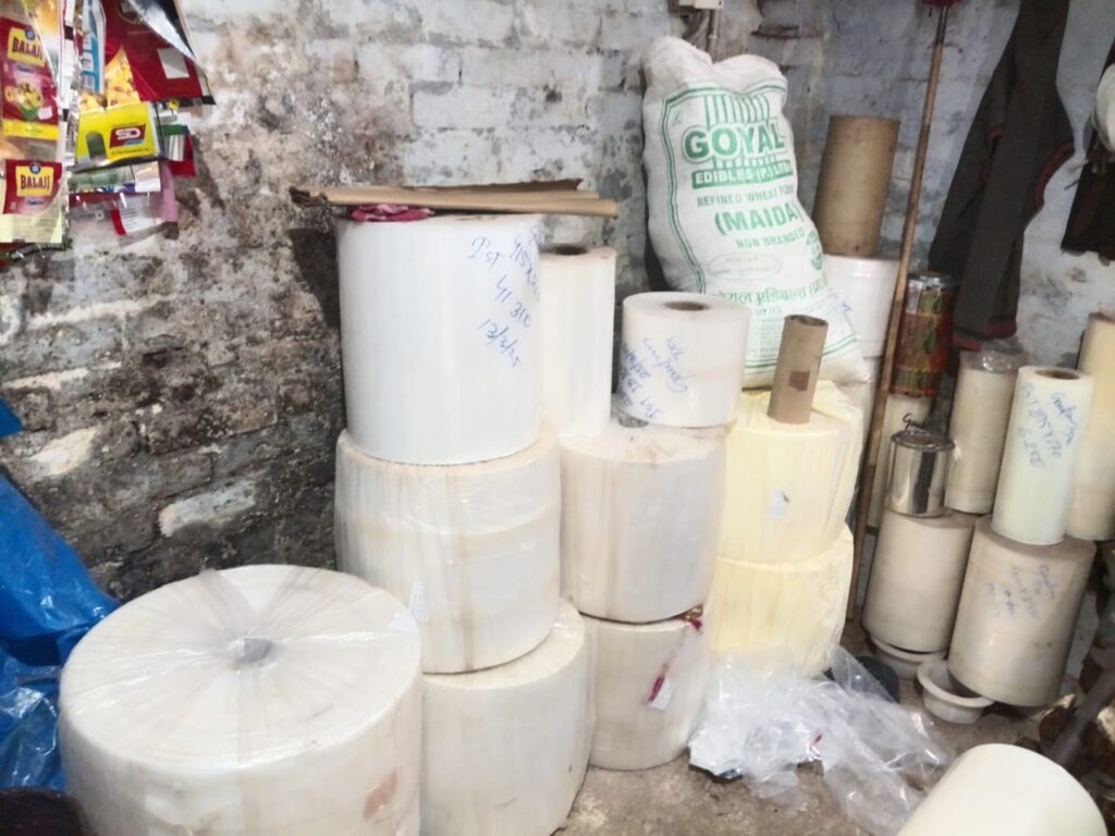 A storage area with various rolls of plastic film and packaging materials stacked against a wall, alongside bags of flour and other items.