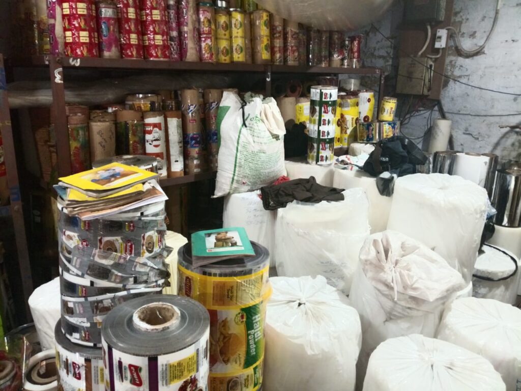 A cluttered storage area filled with various types of packaging materials including plastic rolls, pouches, and bags, showcasing the inventory of a packaging manufacturer.